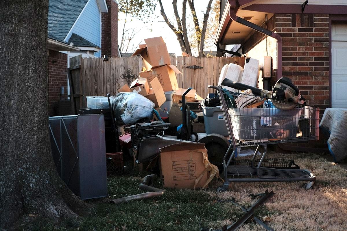 A driveway pile of boxes, a mini fridge, a mattress and yard furniture set out beside a suburban house.