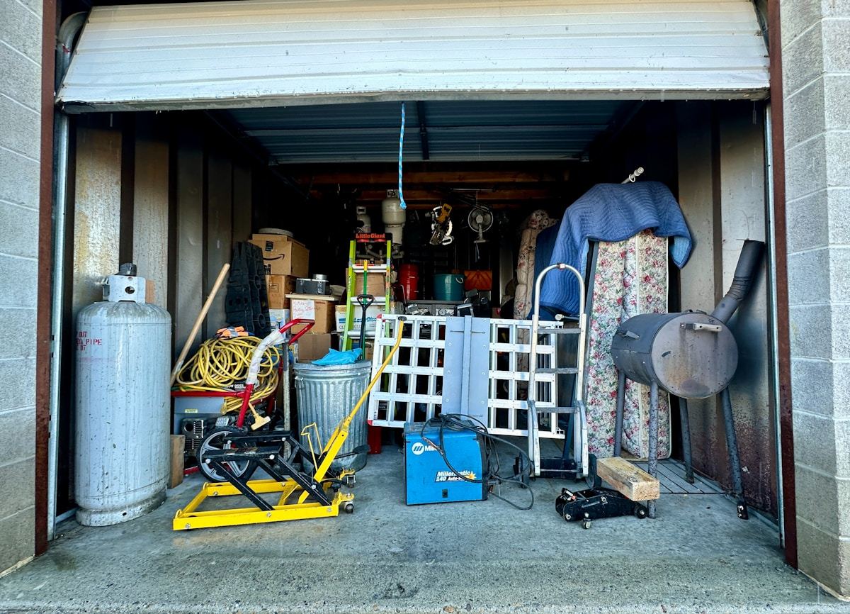 An open garage packed with stored tools, ladders, a mattress, boxes and a propane tank.