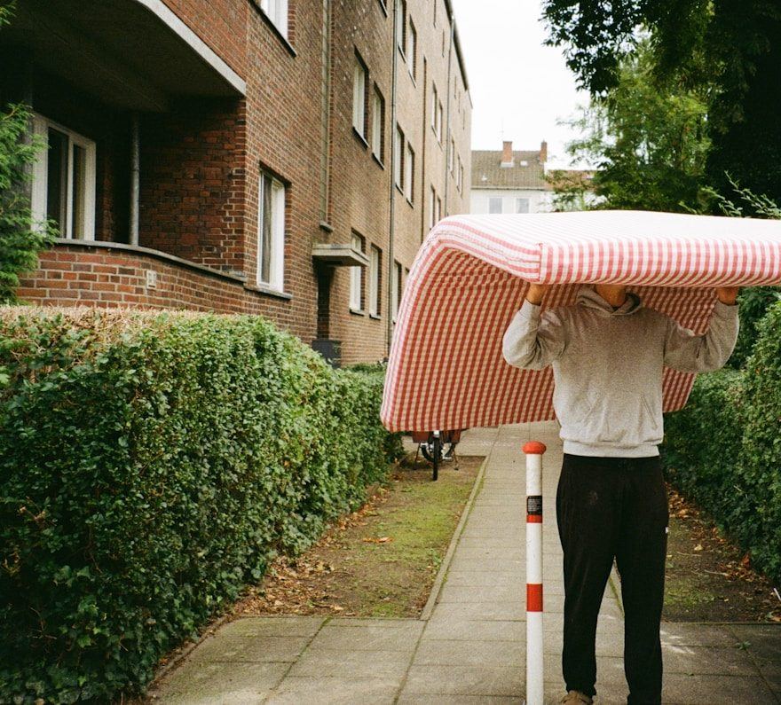 A person carrying a mattress overhead along a residential footpath.